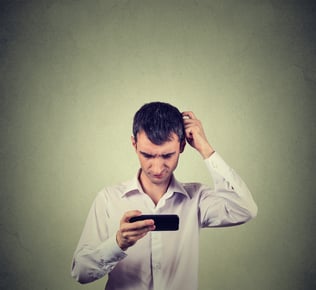 Closeup portrait perplexed young man looking at smart phone seeing bad news or photos with confused emotion on his face isolated on gray wall background. Human reaction, expression