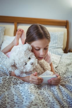 Happy girl lying on bed with a teddy bear and using mobile phone at home