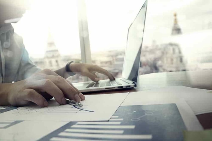 business documents on office table with laptop computer and graph financial diagram and man working in the background-min